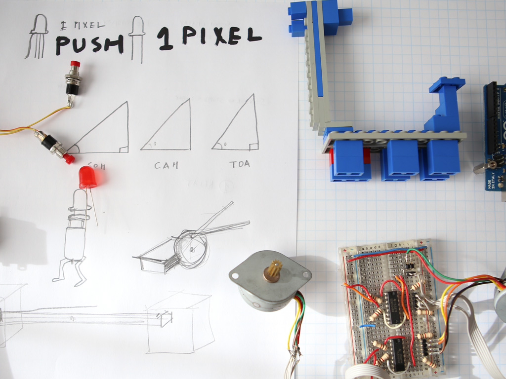 Assortment of tools and toys on a graph paper surface and handwritten sharpie spells out push 1 pixel. Lego, buttons, soldering, and breadboards are strewn about the table.