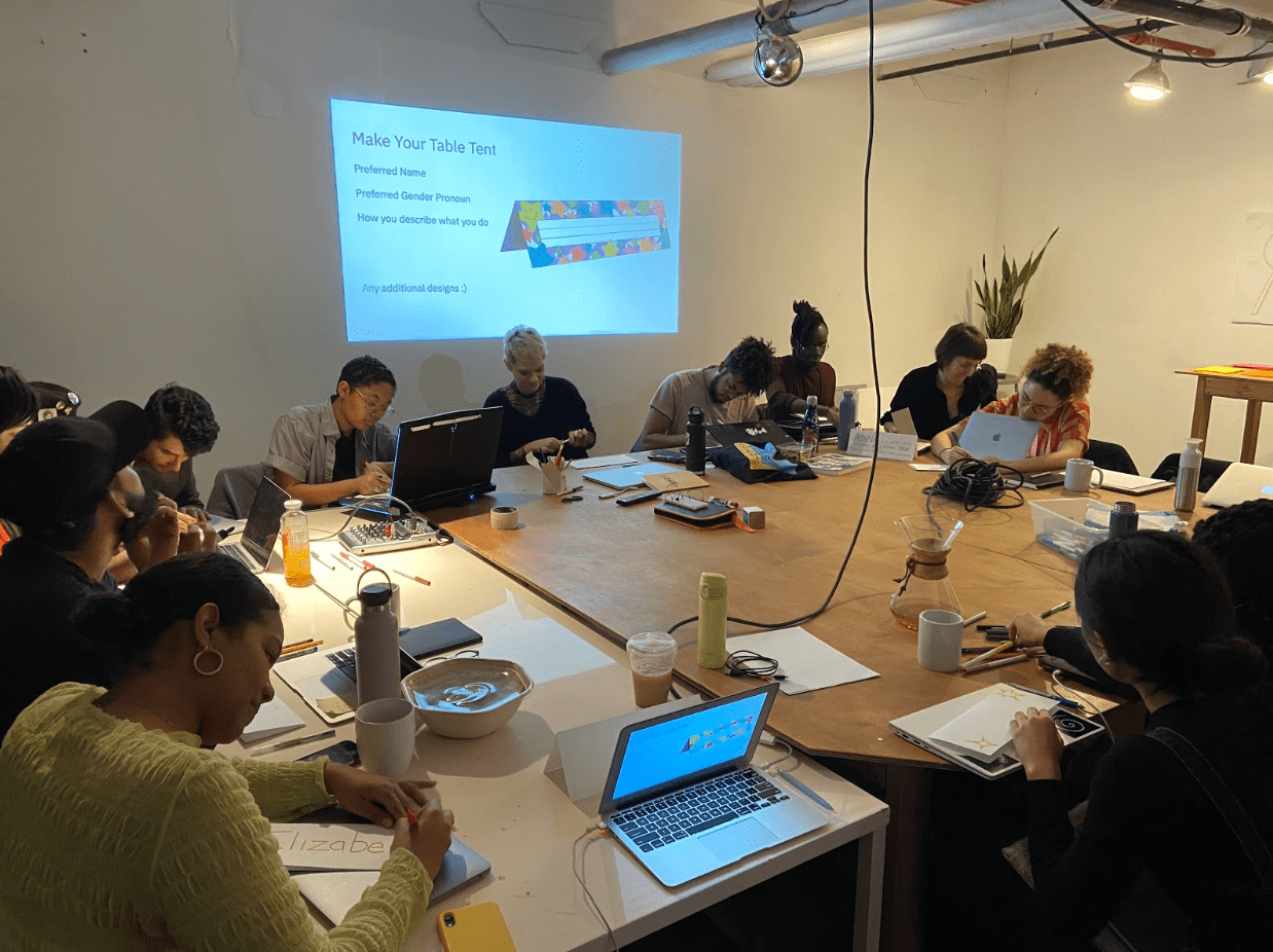 A group of Code Societies students sit around a table drawing on rectangular pieces of paper.